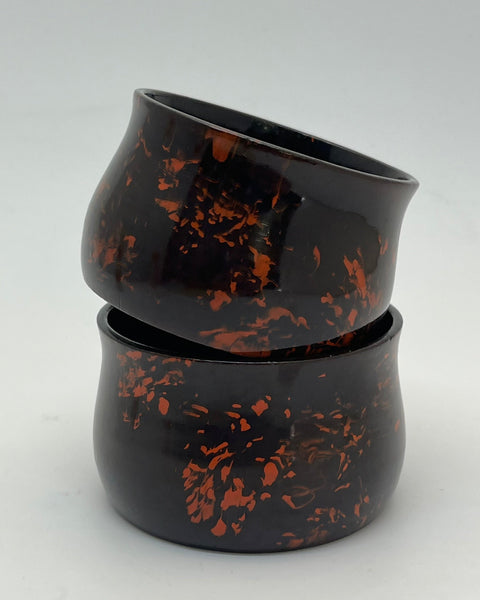 Two stacked glossy black ceramic bowls with orange-brown marbled speckles, photographed on a light gray studio background.