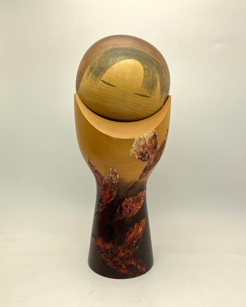 A handcrafted wooden Kokeshi doll by Masao Watanabe a smooth, rounded head in light natural wood, a curved neck in warm tan, and a tapered base decorated with dark brown and reddish-brown marbled wood patterns, set against a plain light gray background.