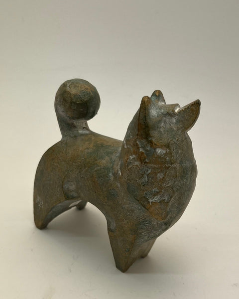 A small sculptural figurine of a dog with a textured, metallic bronze finish, standing on a plain light gray background.