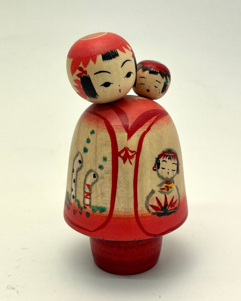 A traditional Japanese wooden kokeshi doll with a red and cream-colored kimono, featuring hand-painted details of children and animals, standing on a red base against a plain white background.