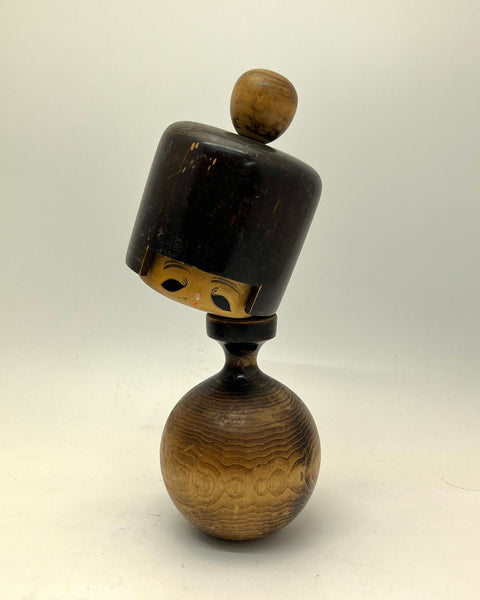 A small carved wooden kokeshi with a rounded brown base, a narrow dark neck, and a tilted dark cap-like top with a round knob, photographed on a plain light background.