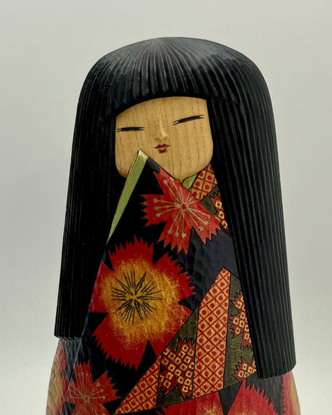 A traditional Japanese kokeshi doll with a black hair bun, painted face, and a patterned kimono in red, orange, and black, 